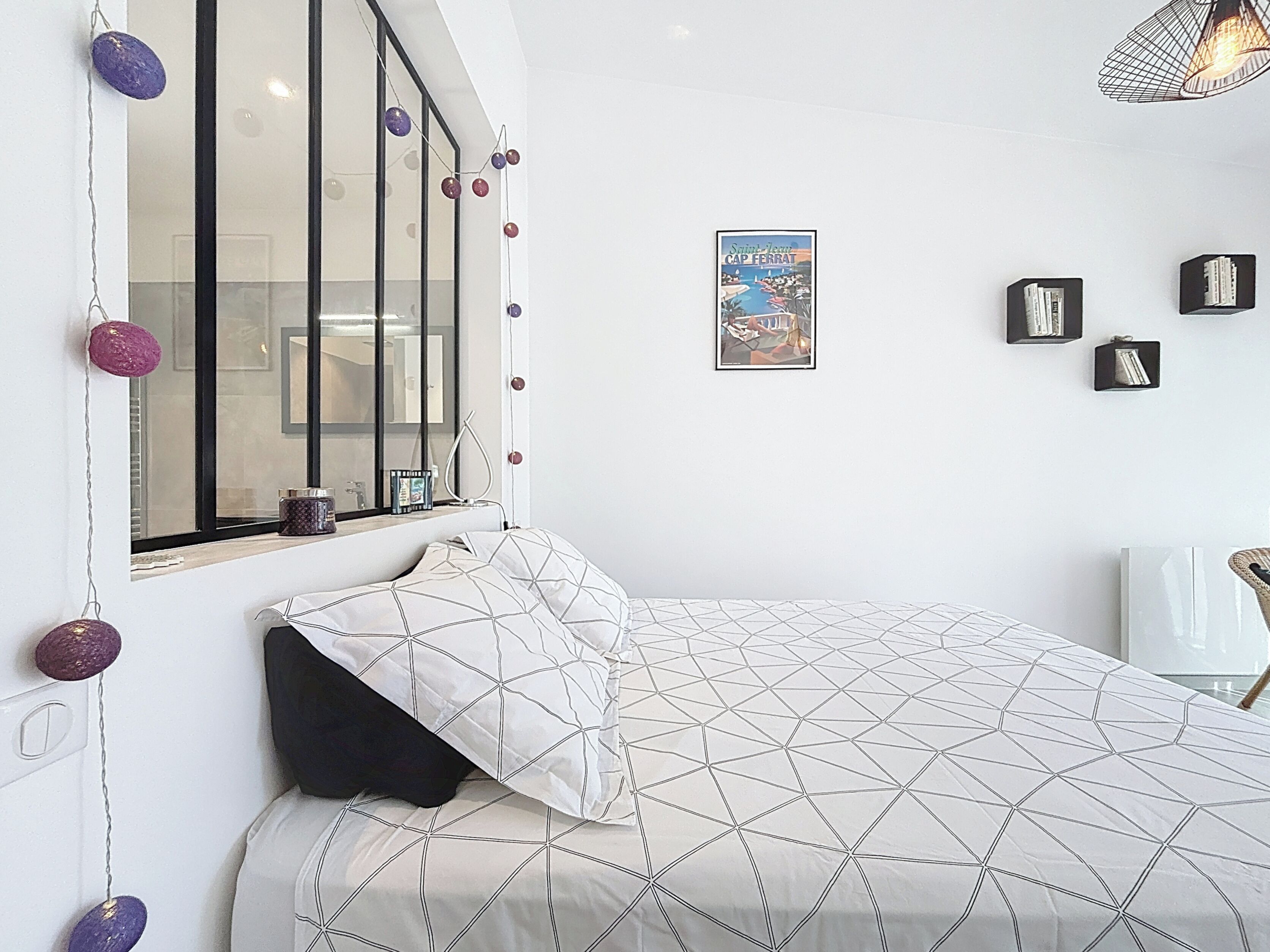 Une chambre à coucher avec un lit, une fenêtre avec des rideaux, une affiche accrochée au mur et une petite table avec une lampe.