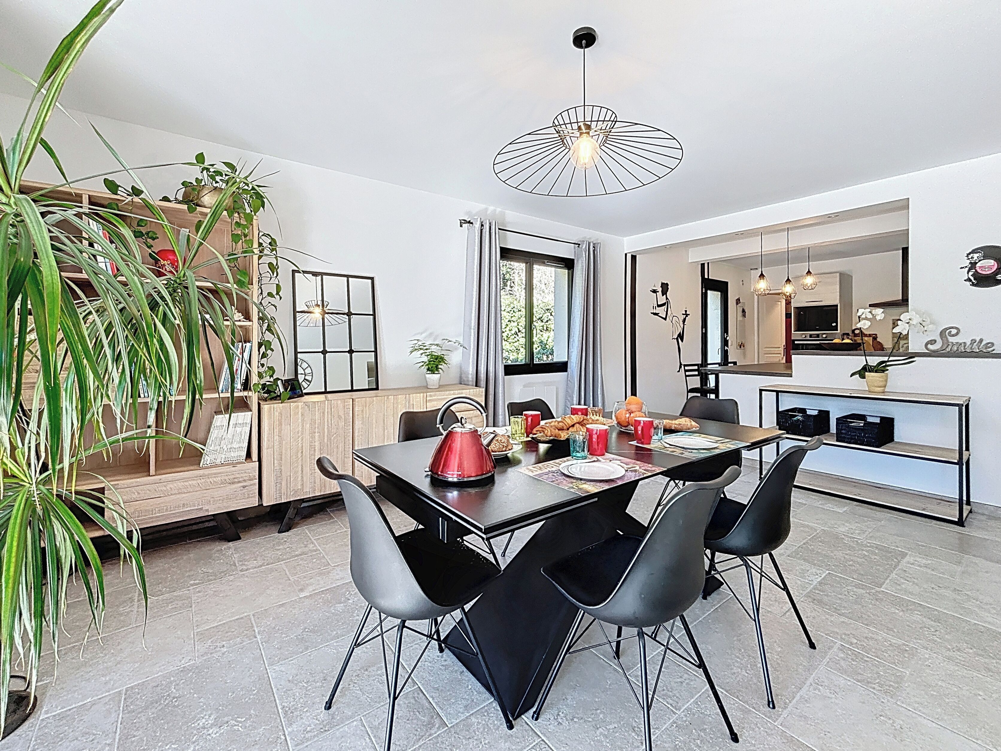 Un espace repas moderne avec une table en bois, des chaises noires et une bouilloire rouge. On y trouve des plantes, un miroir et une vue sur la cuisine.