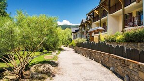 A pathway leading to a row of wooden buildings with balconies, surrounded by greenery and a stone wall.
