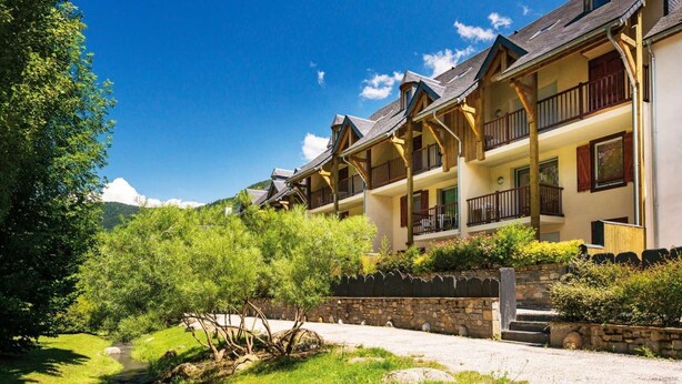 A row of wooden buildings with balconies, a stone wall, and a grassy area with a stream.