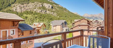 A wooden balcony with a table and chairs, overlooking a mountainous area with buildings.