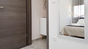 Moderni hotellihuone, jossa on puinen ovi, valkoinen yöpöytä, valkoisella peitteellä varustettu sänky ja suuri peili, joka heijastaa huonetta.