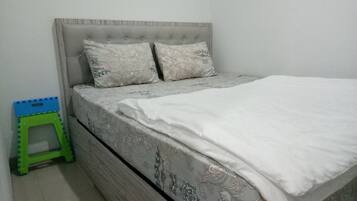 A bed with a grey upholstered headboard and patterned bedding, a green and blue step stool, and a plain white wall.