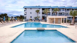 Une piscine entourée de chaises longues et de parasols, elle-même bordée d’un immeuble à plusieurs étages doté de balcons.