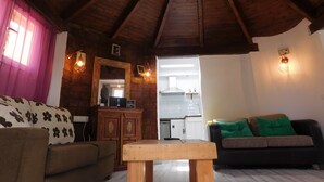 Ein gemütliches Wohnzimmer mit Holzbalkendecke, einer Couch mit gemusterten Kissen, einer schwarzen Couch mit grünen Kissen, einem Holztisch und einem durch einen offenen Durchgang einsehbaren Küchenbereich.