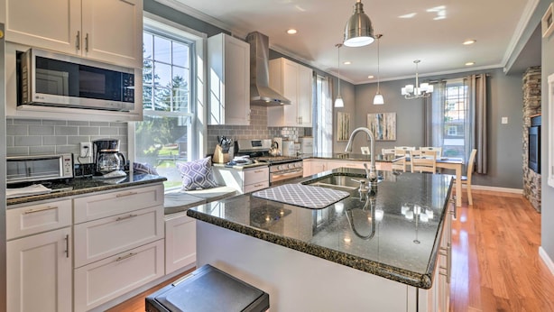 A modern kitchen with white cabinets, a large island with a sink, and a dining area with a table and chairs. - Walk to Parks & Dining: Spacious Ligonier Home! (Ligonier)