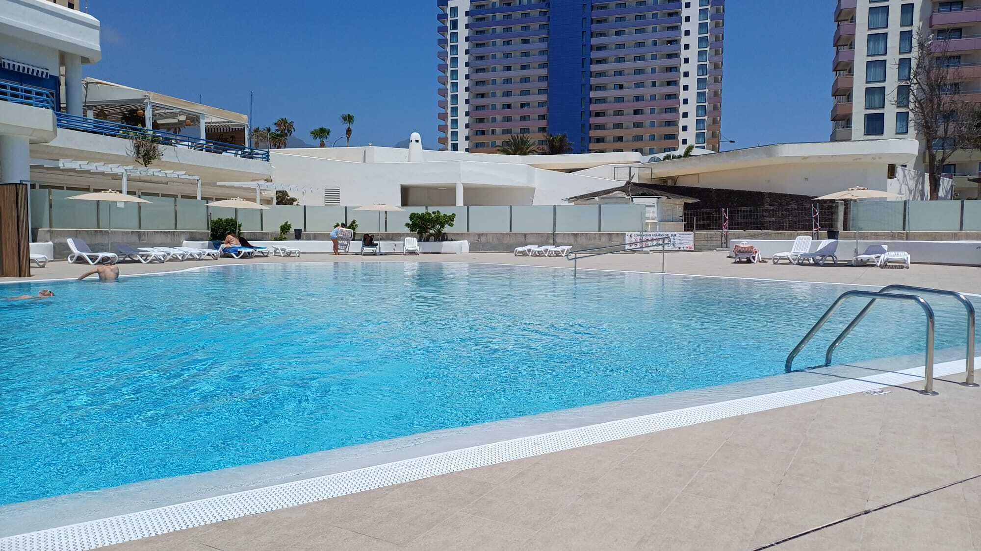Una piscina con sillas de descanso, un puesto de socorrista y edificios altos al fondo.