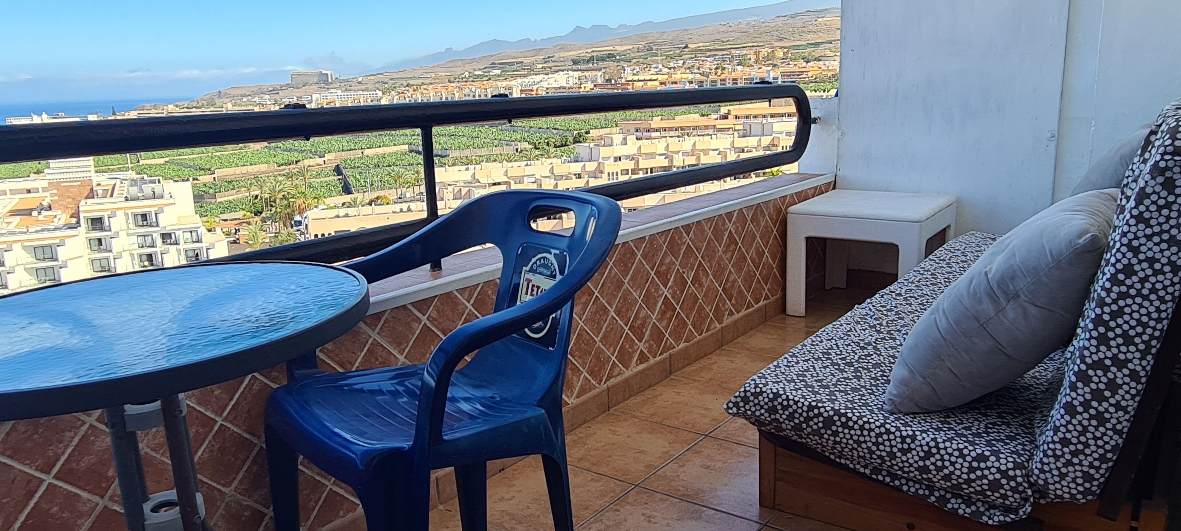 Ein Balkon mit rundem Tisch, blauem Kunststoffstuhl, einem kleinen weißen Hocker und einer gepolsterten Couch mit Kissen.