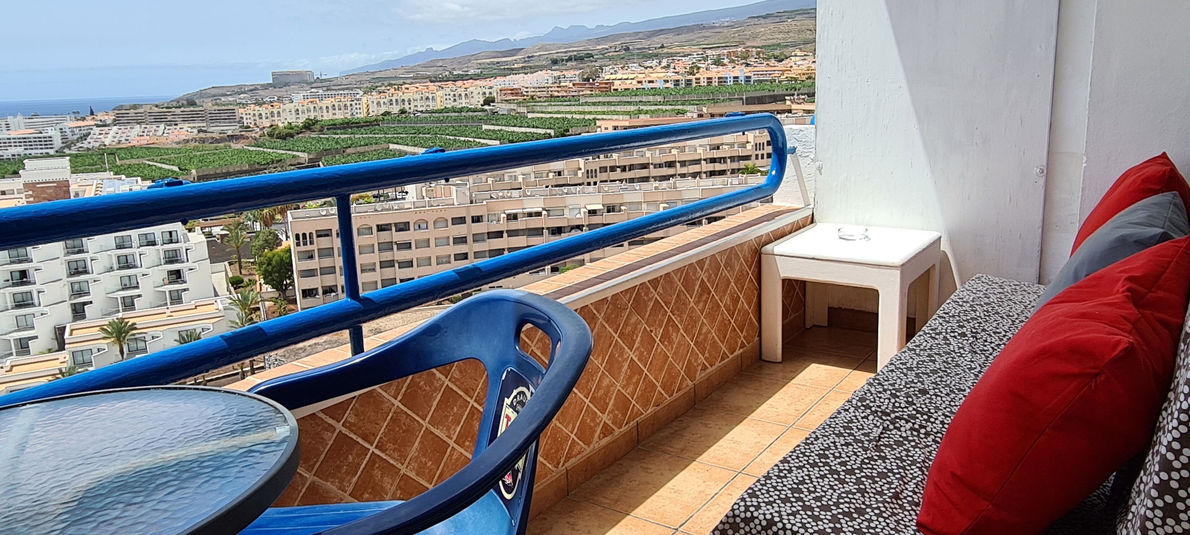 Un balcón con vista a una ciudad costera, una silla azul, una mesita pequeña y un mesita de noche blanco con cojines rojos y grises.