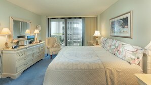 A hotel room with a bed, dresser, chair, and a large window with blinds. - South Seas Tower Two # 810.  Condo with Balcony, Beach, Pool and Fitness Facility. (Marco Island)
