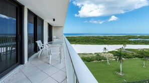 A balcony with a glass door, a small table with chairs, and a view of a beach and ocean.