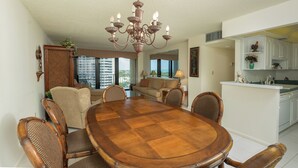 A dining room with a wooden table and chairs, a chandelier, and a view of the city through large windows. - South Seas Tower Two # 810.  Condo with Balcony, Beach, Pool and Fitness Facility. (Marco Island)