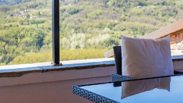 Un balcon avec une table en verre et une chaise, donnant sur un paysage montagneux verdoyant, avec quelques bâtiments visibles au loin.