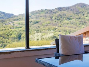 Un balcon avec une table en verre et une chaise, donnant sur un paysage montagneux verdoyant, avec quelques bâtiments visibles au loin.