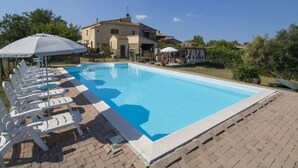 Une piscine entourée de chaises longues et de parasols, elle-même bordée d’une terrasse en briques et d’un bâtiment doté d’un toit en tuiles.