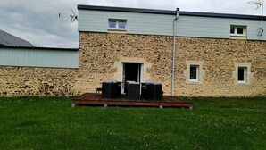 A stone building with a flat roof, a wooden deck with outdoor furniture, and a grassy yard. - GITE: Country house, near the landing beaches, with table football. (Le Molay-Littry)