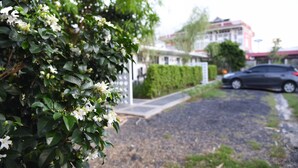 A residential area with a gravel driveway, a car parked on the street, and a building with a red roof in the background. - Snoozy (Phuket)