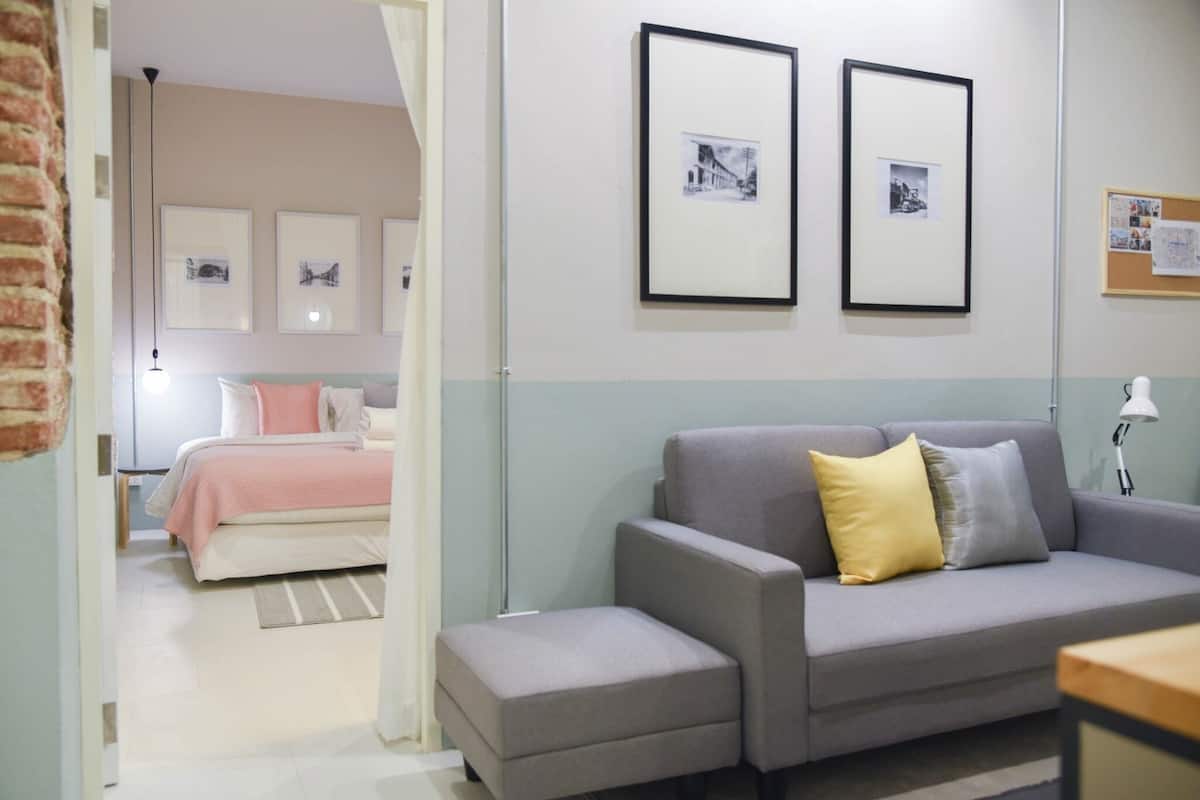 A modern living room with a grey sofa, a small ottoman, and a bed with a pink and white bedspread. There are framed pictures on the wall and a bulletin board to the right.
