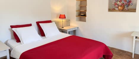 A hotel room with a bed, a bedside table, a lamp, a radiator, a shelf with decorative items, and a painting on the wall.