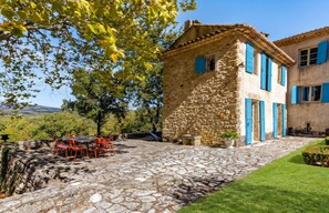 A stone building with blue shutters, a stone patio with a table and chairs, and a view of the countryside.