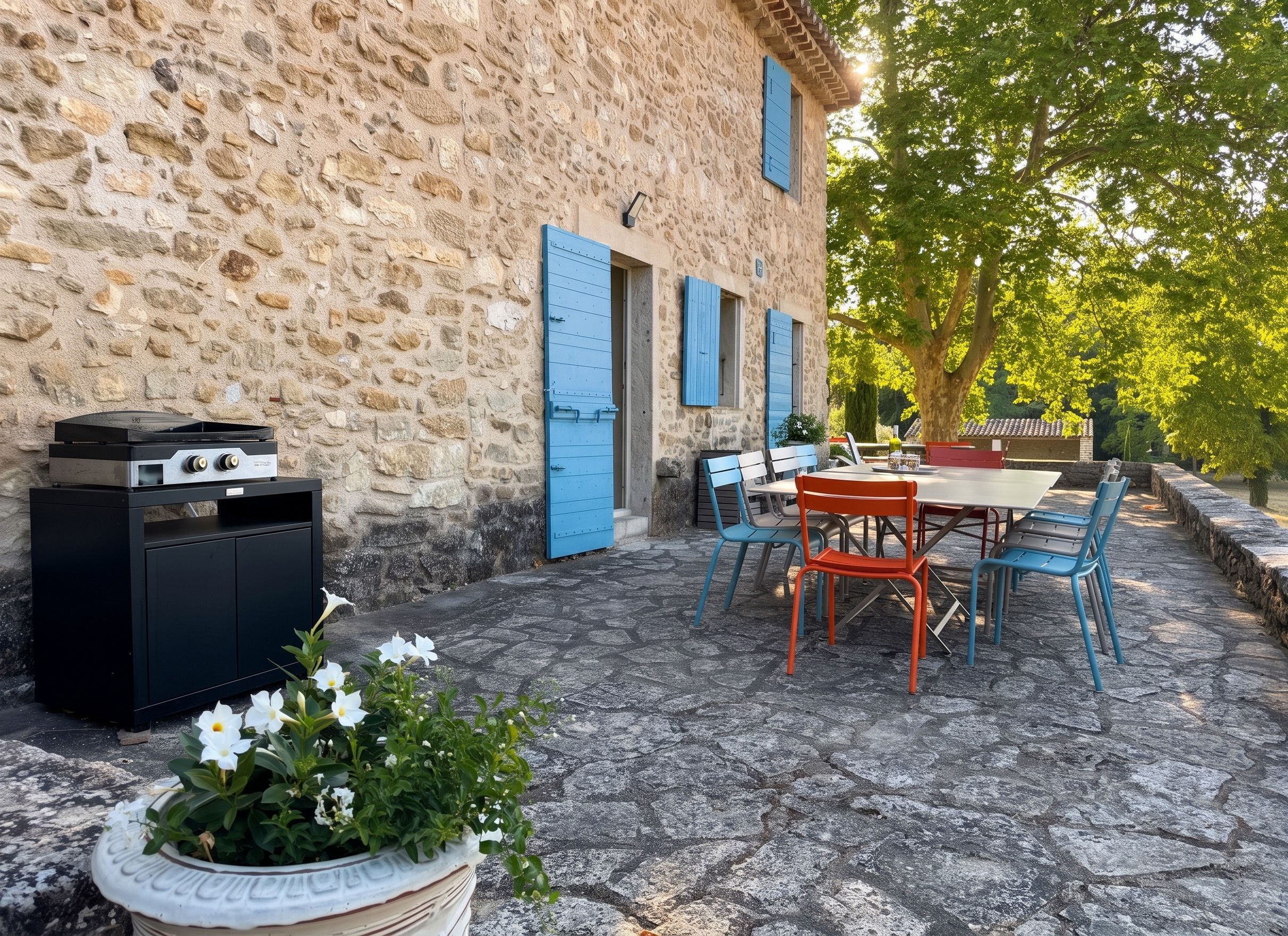 A stone building with a patio area featuring a table and chairs, a grill, and a potted plant with white flowers.