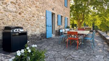 A stone building with a patio area featuring a table and chairs, a grill, and a potted plant with white flowers.