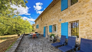 A stone building with blue shutters, a stone patio with outdoor seating, and a view of trees and sky. - Homerez - Villa in Saignon (Saignon)