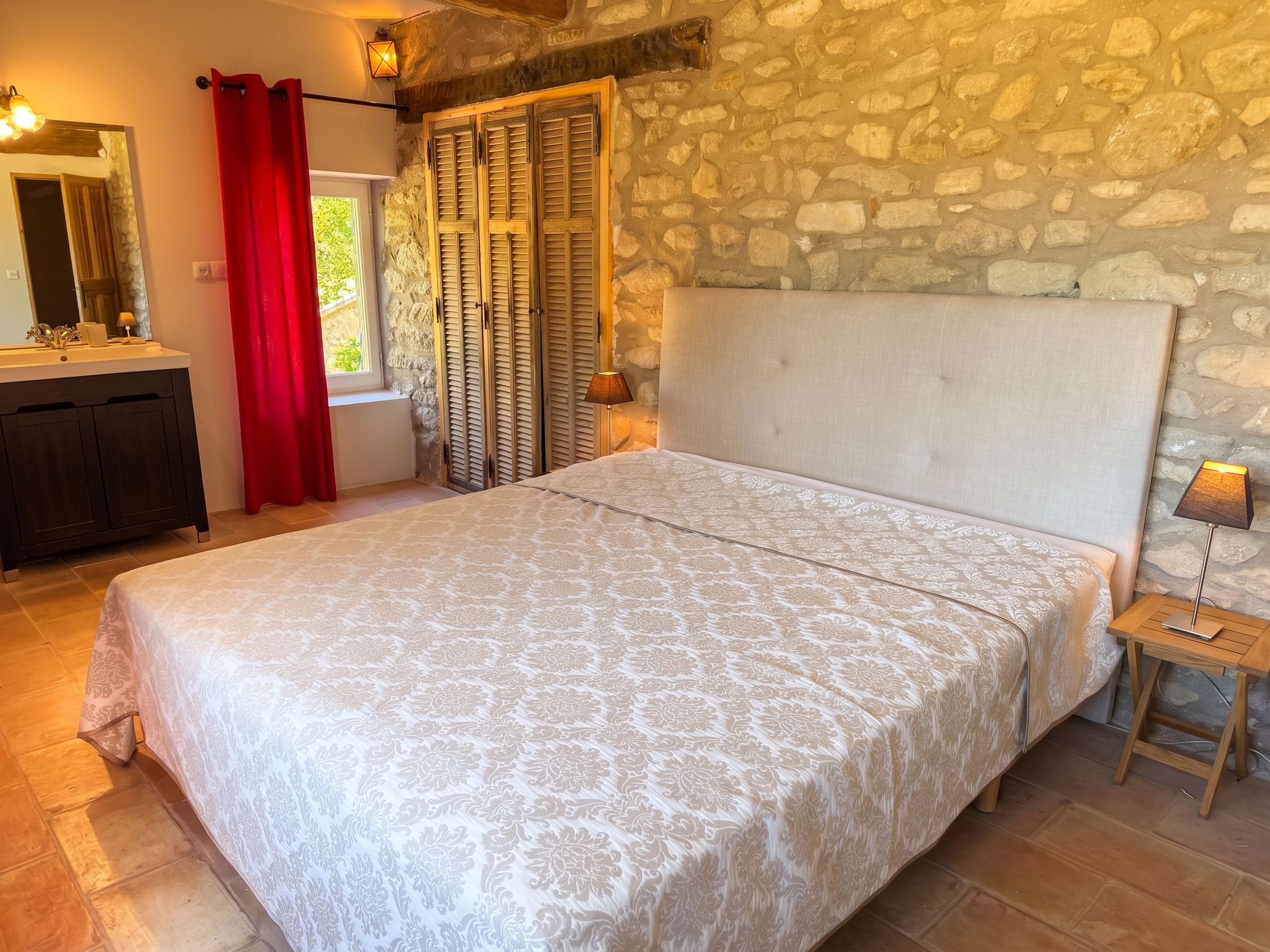 Une chambre avec un mur en pierre, un lit recouvert d’une couvre-lit à motifs, une table de chevet avec une lampe et une fenêtre avec des volets.