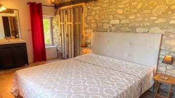 Une chambre avec un mur en pierre, un lit recouvert d’une couvre-lit à motifs, une table de chevet avec une lampe et une fenêtre avec des volets.