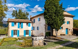 Un edificio de piedra tradicional con contraventanas azules y techo de tejas, rodeado de vegetación y un cielo despejado.