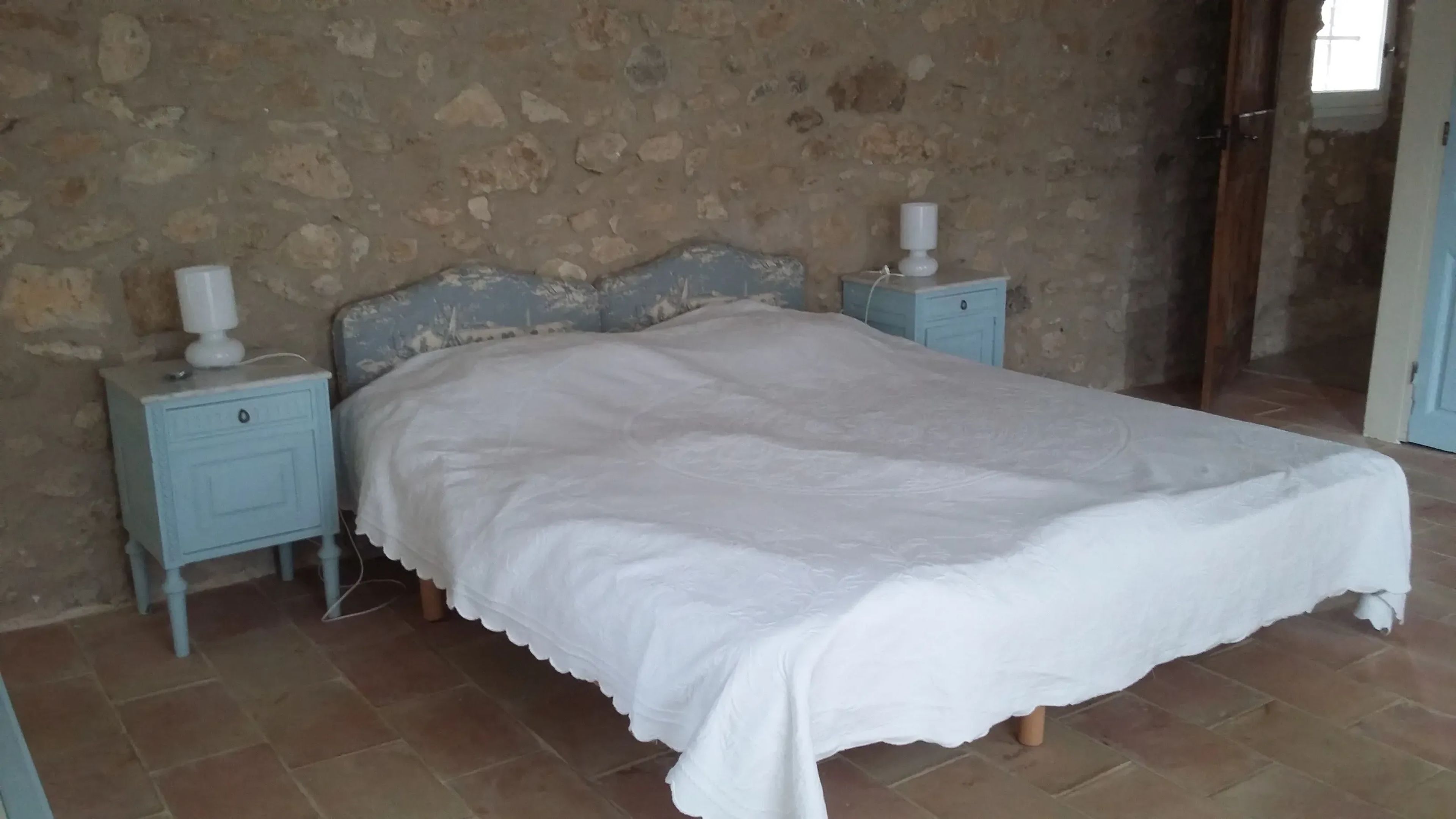 Une chambre avec un mur en pierre, un lit recouvert d’une courtepointe blanche, deux tables de chevet avec des lampes et un plancher carrelé.