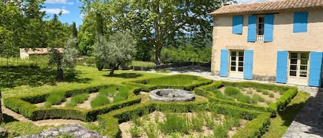 A traditional building with blue shutters, a well-maintained garden with geometric hedges, and a clear blue sky.