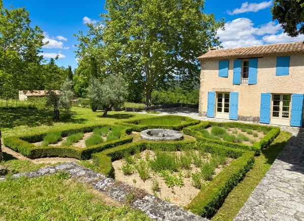 A traditional building with blue shutters, a well-maintained garden with geometric hedges, and a clear blue sky.