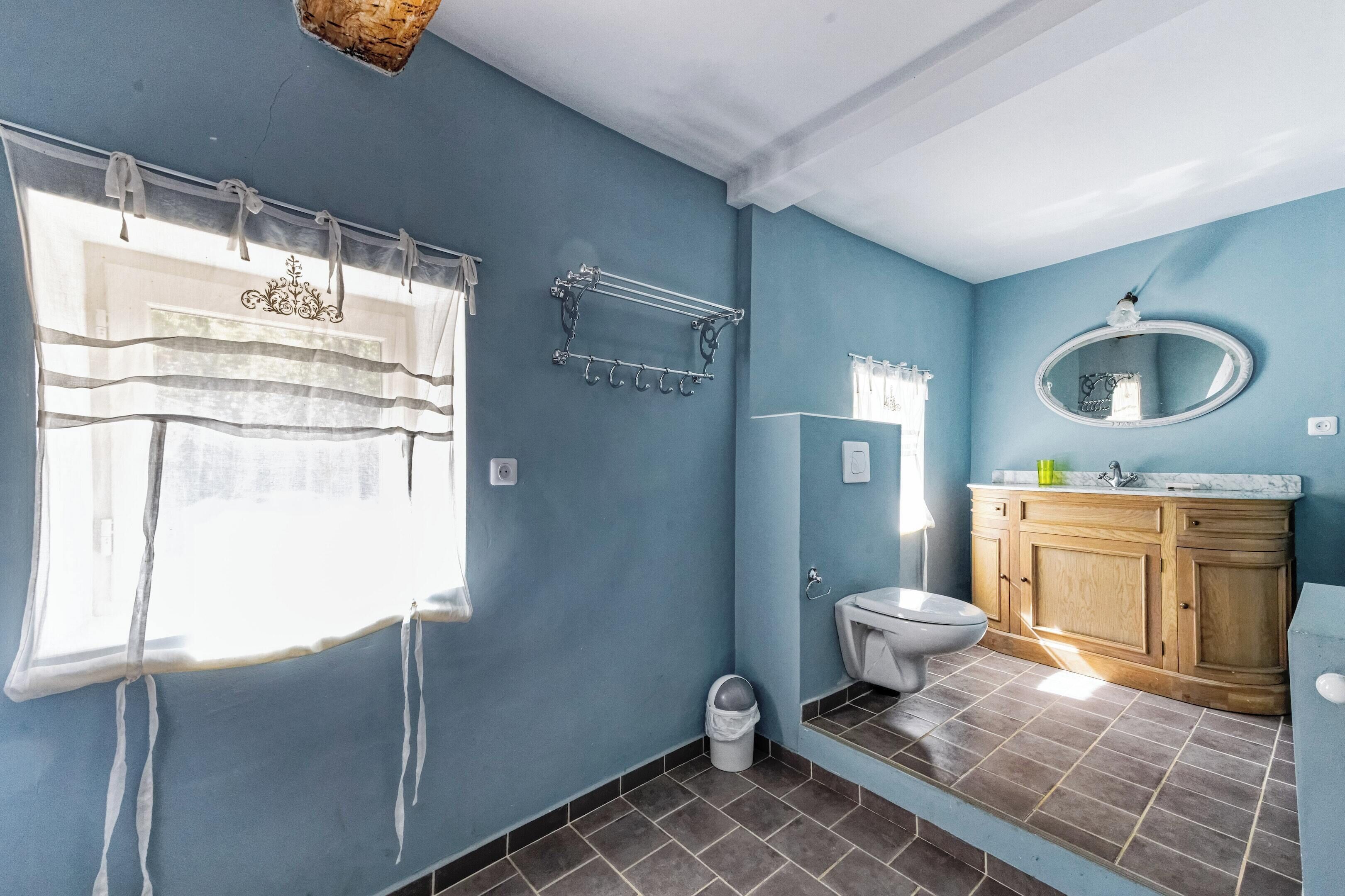 Une salle de bain avec un mur bleu, une fenêtre avec un rideau, des toilettes, un lavabo avec un miroir et un meuble en bois.