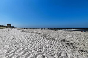 Una playa arenosa con cielo despejado, algunas personas y edificios al fondo.