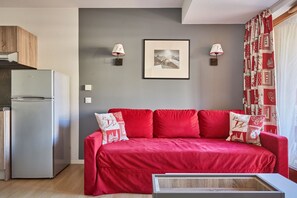A compact living space with a kitchenette, a red sofa with patterned cushions, two wall-mounted lamps, and a framed picture on the wall. - Appart de Montagne Cosy | Service de Navette ! (Serre Chevalier)