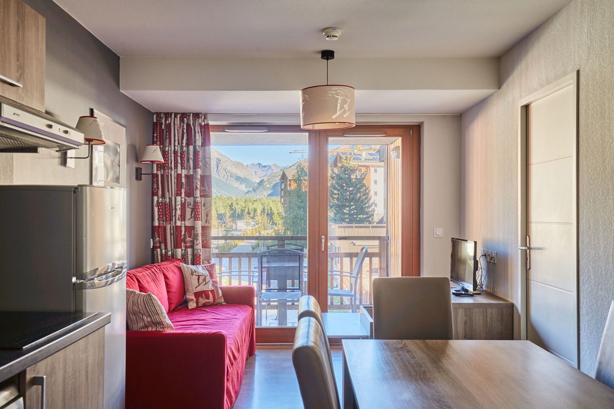 A modern kitchen with a red sofa, dining table, and a view of mountains through the balcony.