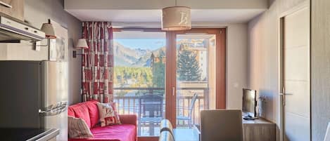 A modern kitchen with a red sofa, dining table, and a view of mountains through the balcony.