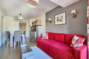 A modern living room with a red sofa, a dining area with chairs, a kitchen with a refrigerator, and a hallway leading to another room.