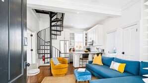 A modern living room with a blue sofa, yellow armchair, and a spiral staircase leading to a kitchen. - 8C at Saint Philip Square, One Block to King, Bright Enchanting Top Floor Apt (Charleston)