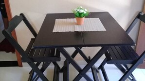 Ein kleiner, quadratischer Tisch mit dunkler Oberfläche, begleitet von vier schwarzen Stühlen schlichten Designs. Auf dem Tisch steht ein kleiner Topfpflanze mit grünen Blättern, und auf der Tischplatte liegt eine hellfarbene Unterlage.