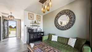 Una sala de estar con un sofá verde, una mesa de centro de madera y un espejo grande y decorativo. Se observa un mueble negro con estantes y un candelabro colgante.