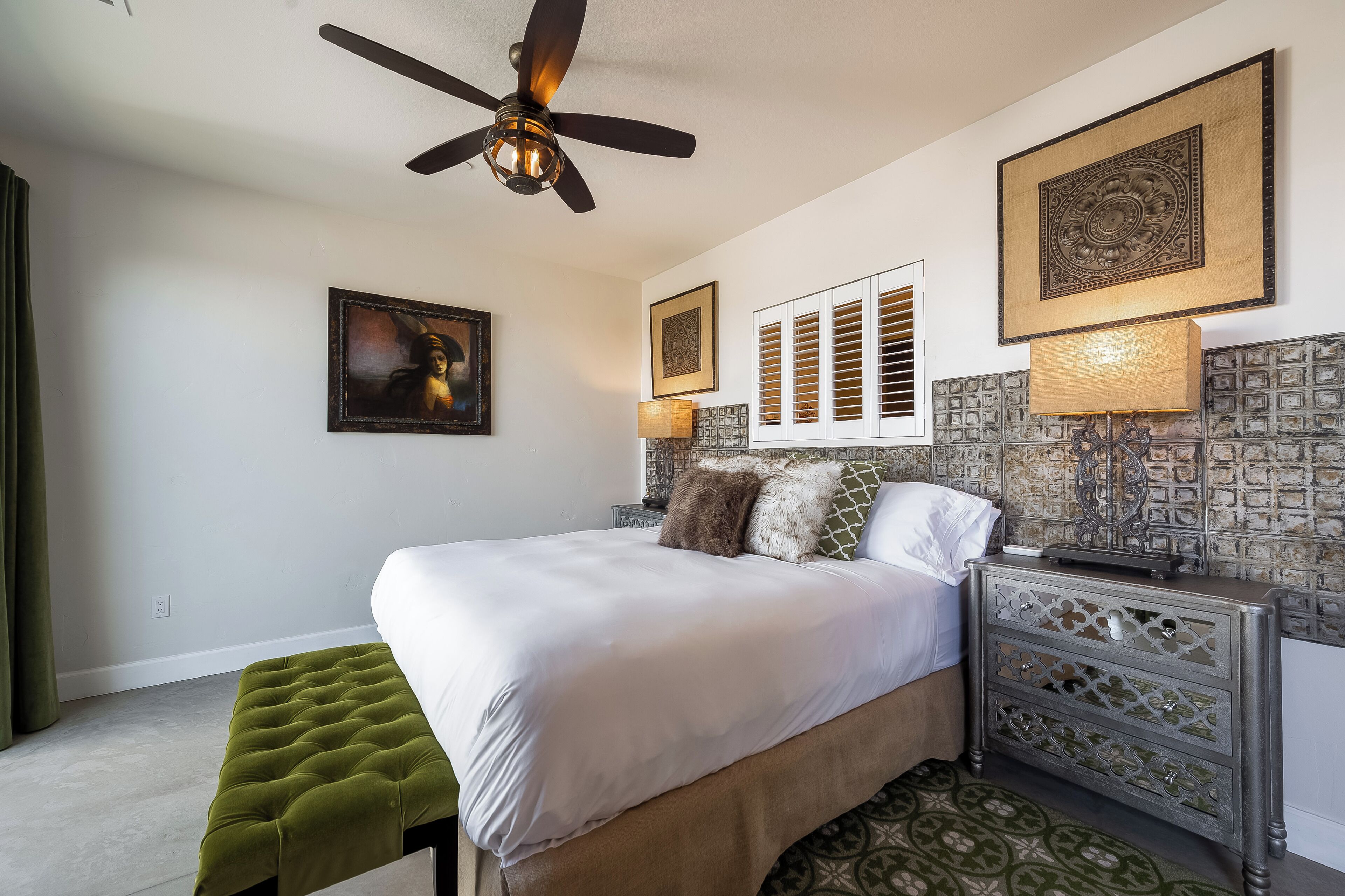 A bedroom with a bed, a green ottoman, a ceiling fan, and decorative wall panels.