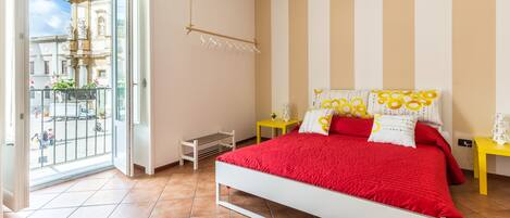 A bedroom with a white bed, red bedding, and a view of a cityscape through a large window.