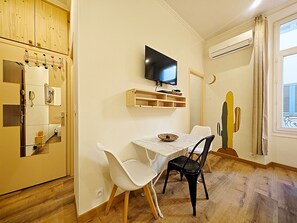 A compact living space with a dining area, a TV mounted on the wall, a wooden cabinet with a phone, and a window with curtains. - Mediterranean studio (Cannes)