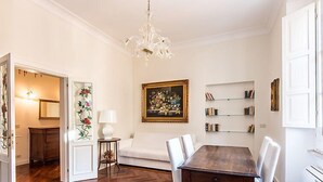 A dining area with a wooden table and white chairs, a chandelier, a painting, and a bookshelf. - Apartment Silla by Interhome (Roma: Vaticano)
