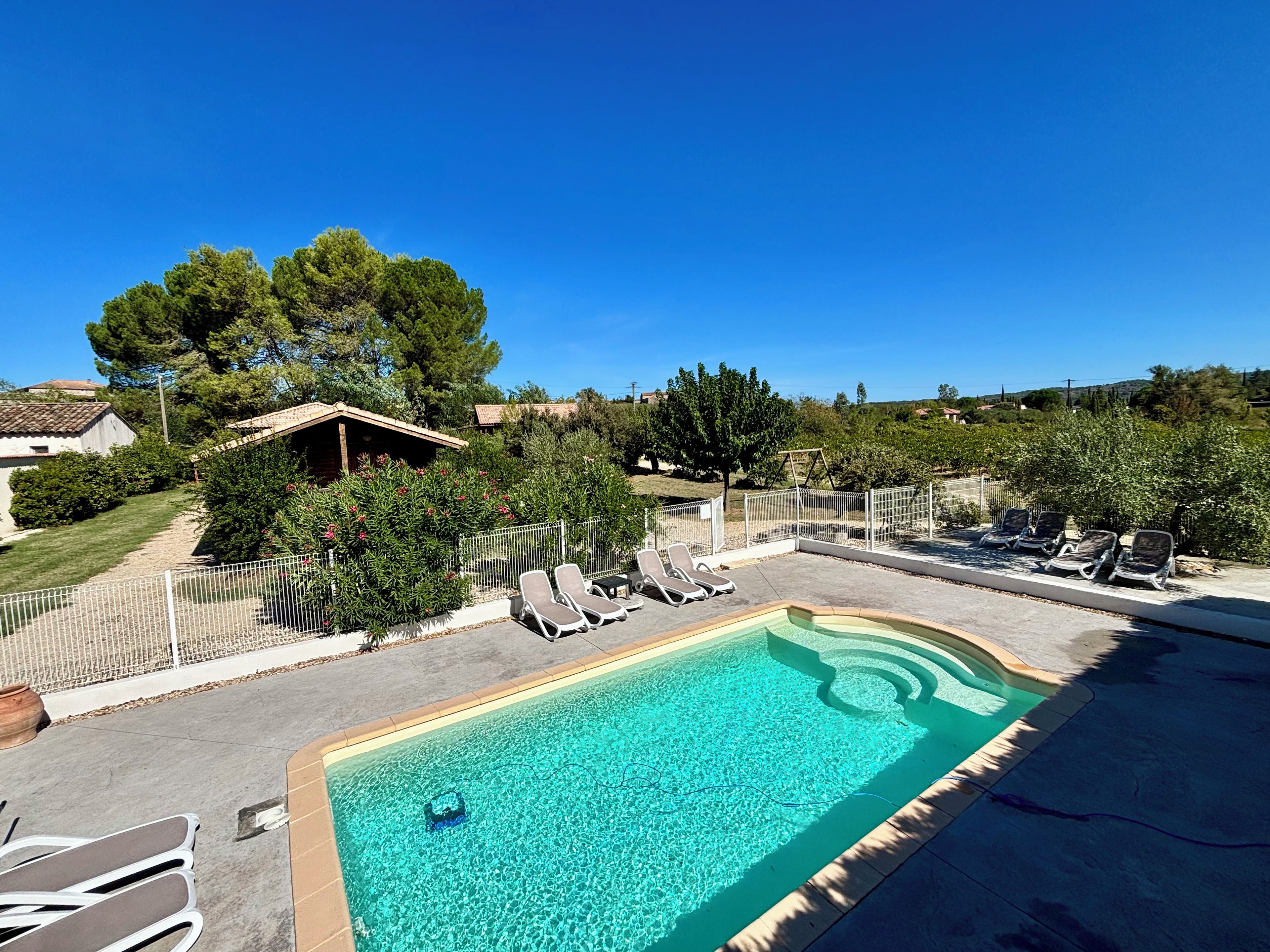Une piscine dotée de chaises longues et d’une zone délimitée par une clôture, le tout entouré d’arbres et d’un bâtiment.