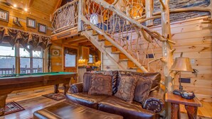 A cozy log cabin interior with a pool table, leather sofa, and a wooden staircase.