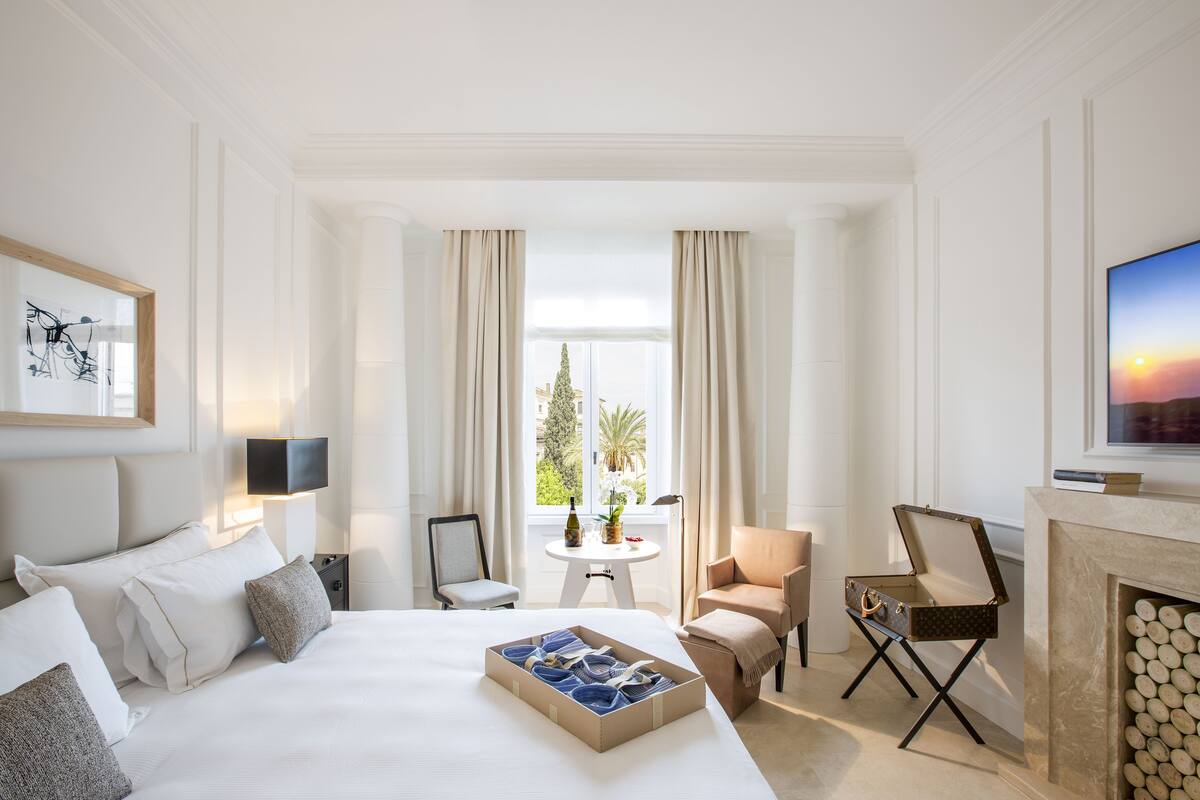 A hotel room with a large bed, a small table with a bottle and flowers, a chair, a television, and a fireplace.