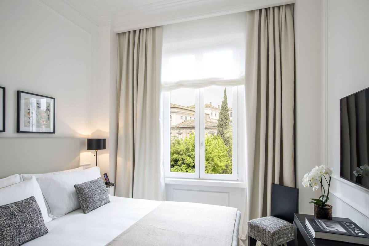 A hotel room with a bed, a bedside table with a lamp, a chair, a window with curtains, and a view of greenery and buildings outside.
